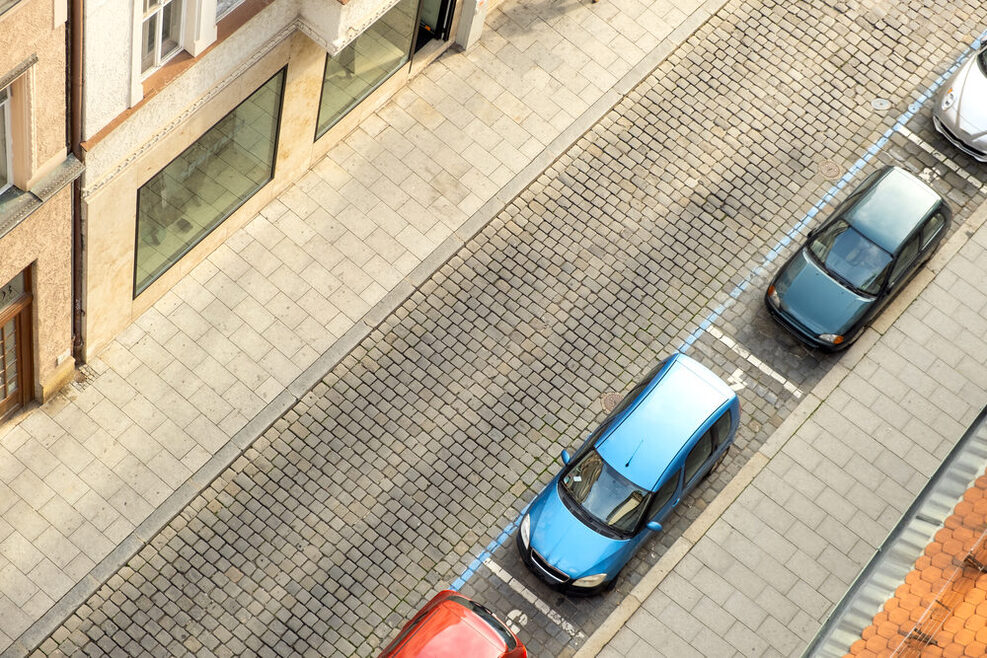 Blick von oben auf eine einspurige Straße an deren rechter Seite Autos in Reihe parken. Links sind Häuserfronten zu erkennen.