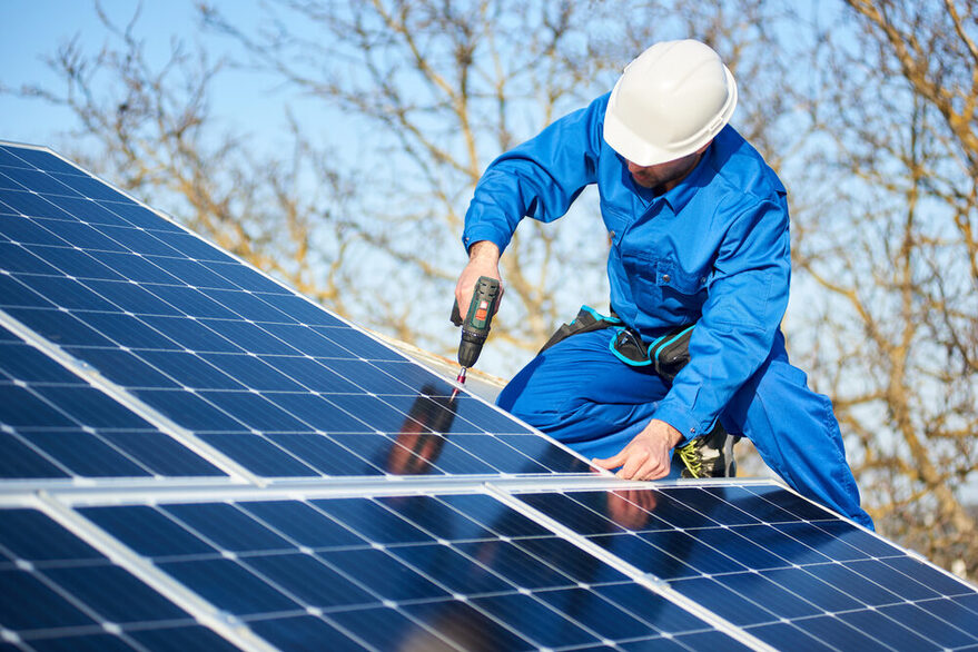Auf dem Dach eines Hauses befestigt ein Bauarbeiter in blauer Arbeitskleidung Solarpanele.