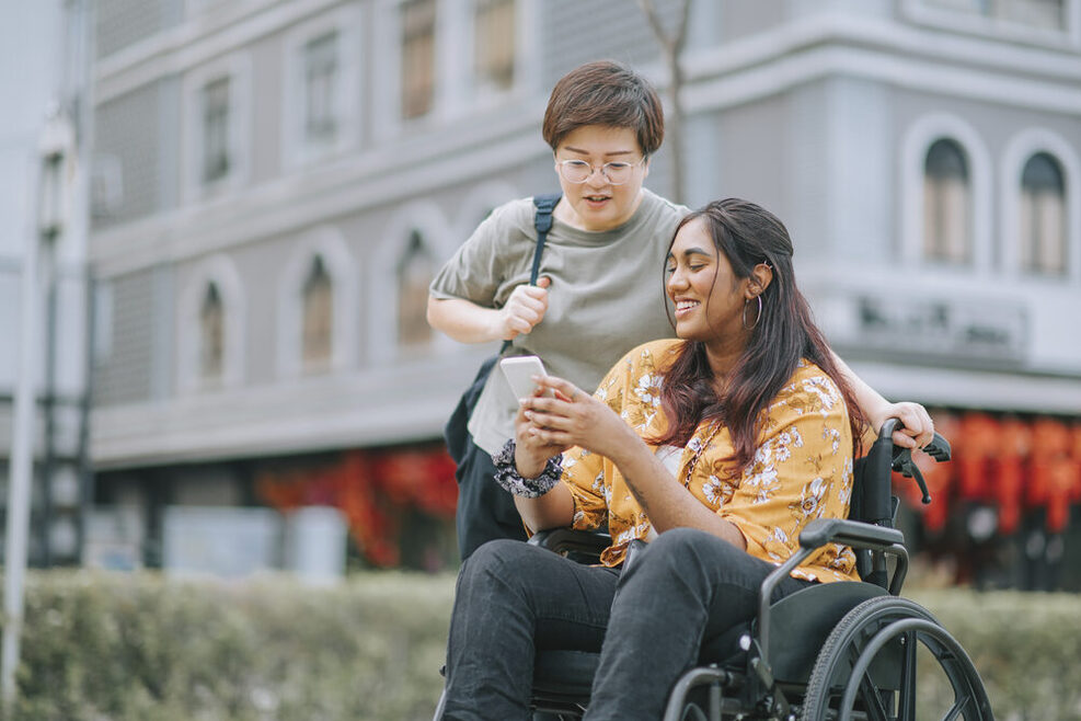 Eine Rollstuhlfahrerin siht lächelnd in ihr Handy während iene junge Frau neben ihr steht.