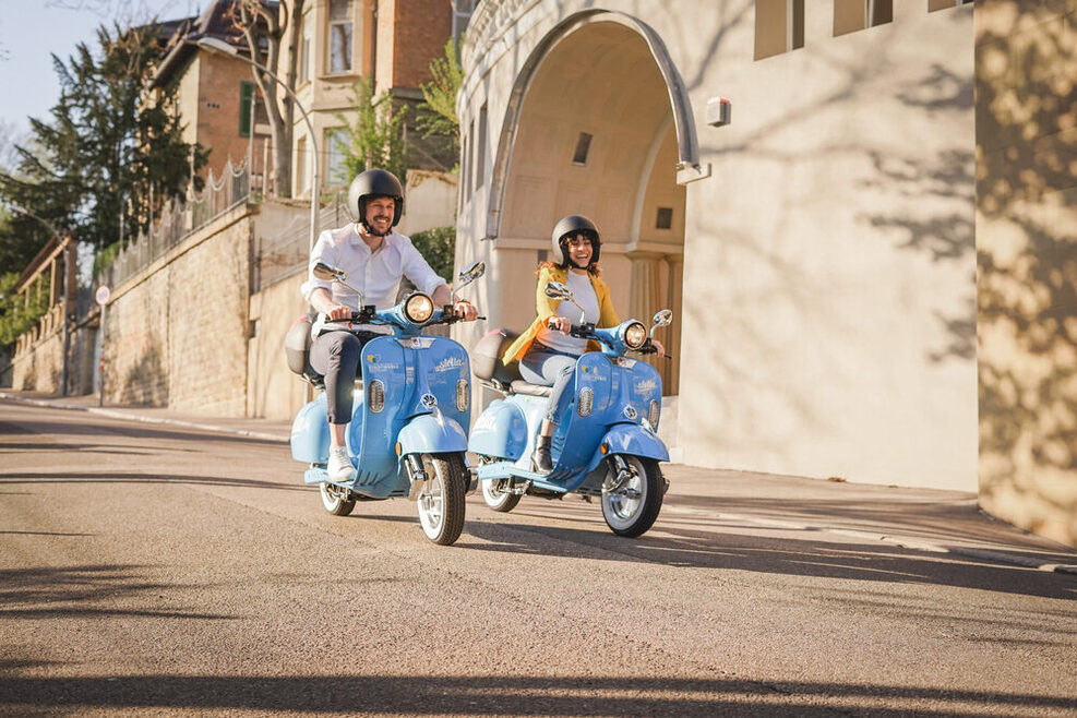 Ein Mann und eine Frau fahren auf einem blauen E-Roller eine Straße entlang. Im Hintergrund sind Stadtvillen zu sehen. Es ist Frühling.