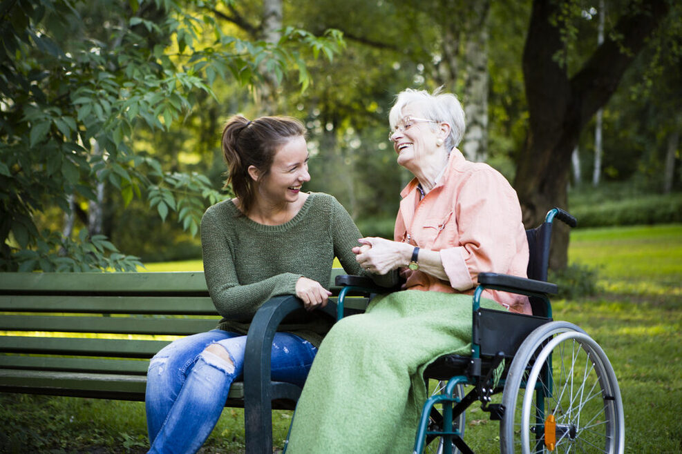 Eine ältere Frau in einem Rollstuhl neben einer auf einer Bank sitzenden jungen Frau im Park. Sie lachen gemeinsam.