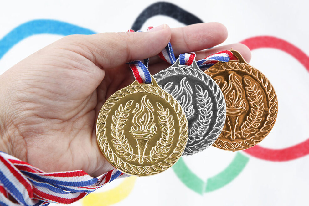 Ein Mann hält drei Medaillen (gold, silber, bronze) in der Hand. Im Hintergrund sind die olympischen Ringe zu sehen.