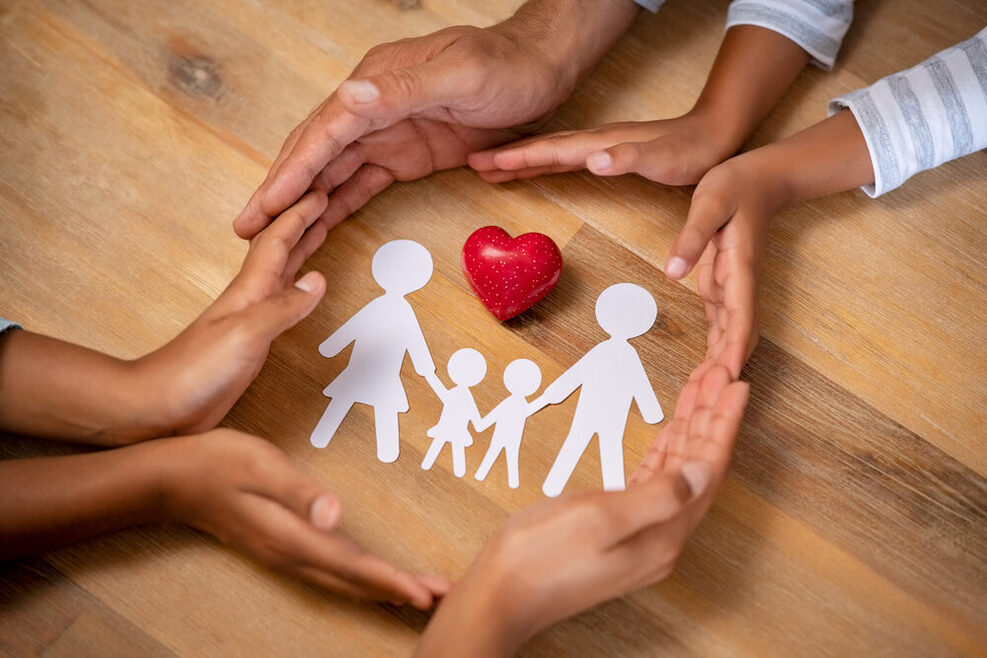 Hände von vier Familienmitgliedern liegen schützend um ein rotes Herz