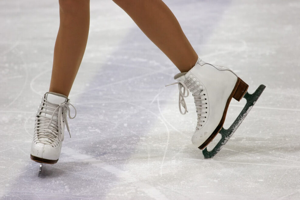 das Bild zeigt Beine und Schlittschuhe einer Eiskunstläuferin, die auf dem Eis steht.