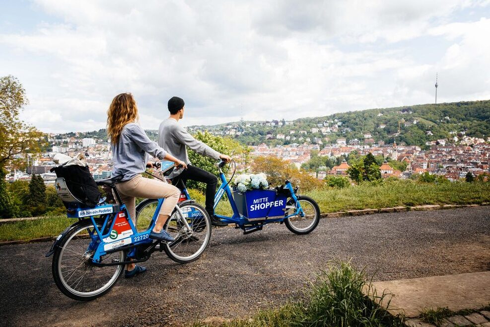 Ein Mann und eine Frau sitzen jeweils auf einem E-Lastenrad und schauen von einer Anhöhe auf Stuttgart und den Fernsehturm.