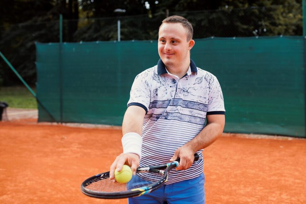 Ein Jugendlicher mit Down-Syndrom hält einen Tennisschläger auf dem ein gelber Tennisball liegt.