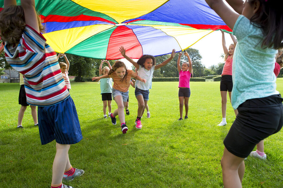 Kinder spielen draußen auf einer Wiese. Zwei Mädchen rennen unter einer Plane durch, die andere Kinder in die Luft halten.