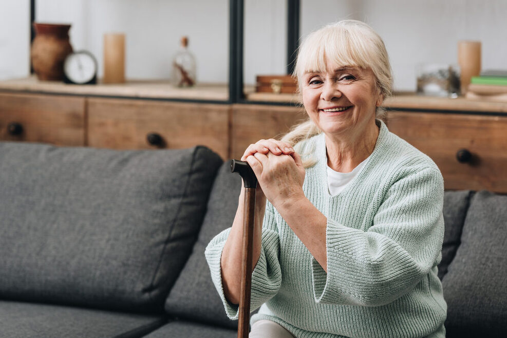 Eine ältere lächelnde Frau sitzt auf einem grauen Sofa, in ihrer Hand hält sie eine Gehhilfe.