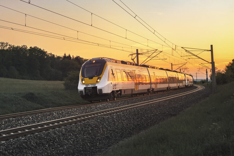 Ein Zug fährt auf einer Bahnstrecke. Links sind eine Wiese und Bäume zu sehen, im Hintergrund ein Sonnenuntergang.