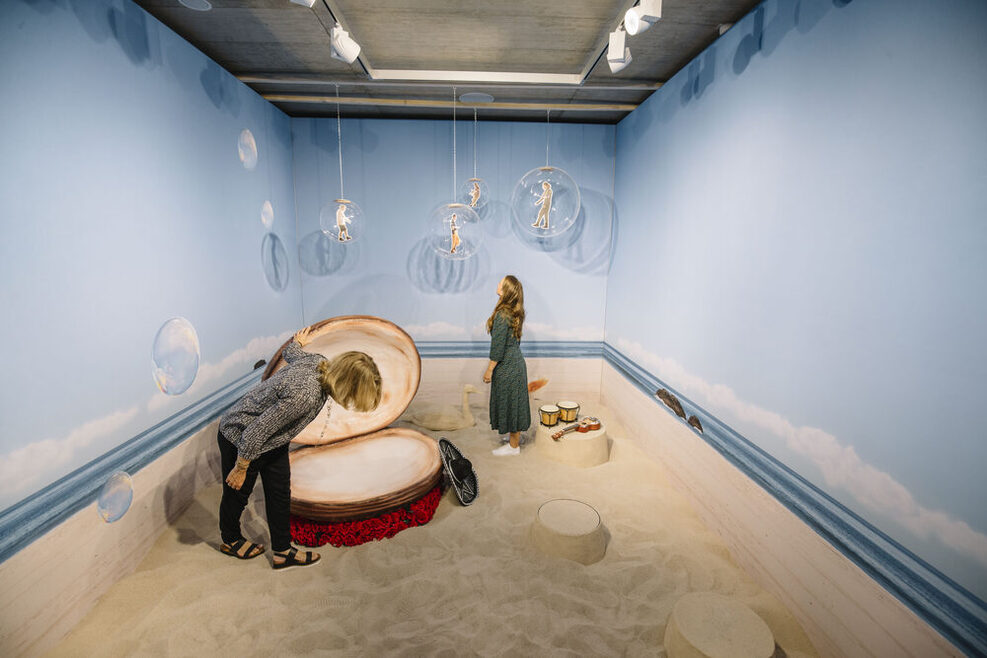 Zwei Jugendliche stehen in einem Ausstellungsraum. Auf dem Boden ist Sand, die Wände sind mit einem Himmel bemalt und von der Decke hängen große Seifenblasen mit Figuren darin. Ein Mädchen schaut in eine übergroße Muschel neben der ein Sombrero liegt. Ein anderes schaut sich eine der Seifenblasen an.
