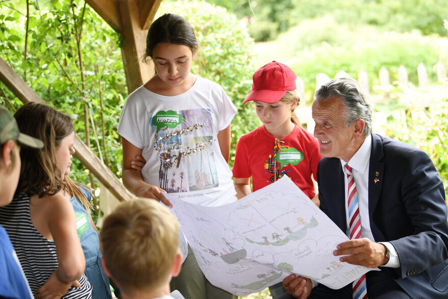 Kinder unterhalten sich mit Oberbürgermeister Dr. Frank Nopper, der eine Planzeichnung von Kindern in den Händen hält und rechts im Bild steht. Im Hintergrund sind Bäume und Grün zu erkennen.