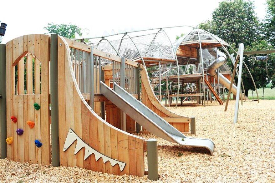 Dino‐Spielplatz aus Holz mi sieben Rutschen, verschiedenen Klettermöglichkeiten und mehrere Schaukeln.