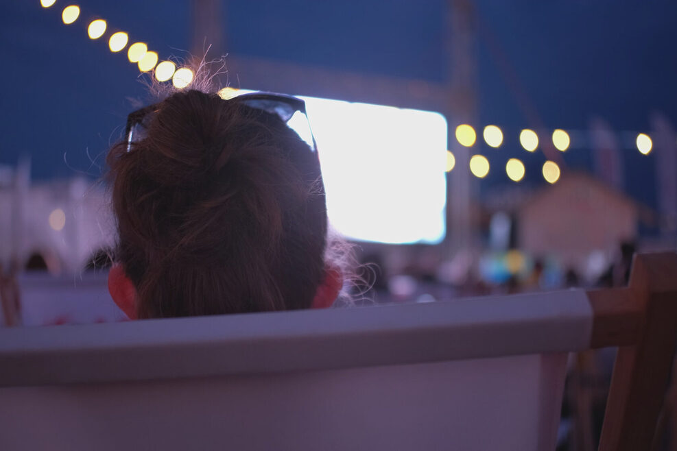Zu sehen ist der Hinterkopf einer Frau, die bei einem Open-Air-Kino auf die Leinwand schaut.