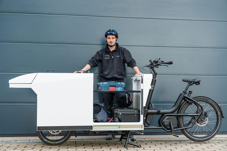 Ein Mann steht mit Fahrradhelm hinter einem Lastenrad, dessen Container vor dem Lenker geöffnet ist. Darin sind Kisten und Taschen verstaut.
