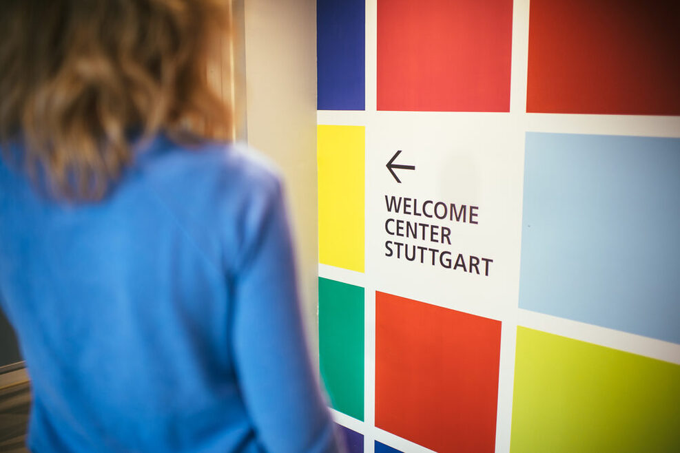 Eine Frau steht vor einer Wand mit bunten Kacheln, auf der Wand steht: Welcome Center Stuttgart