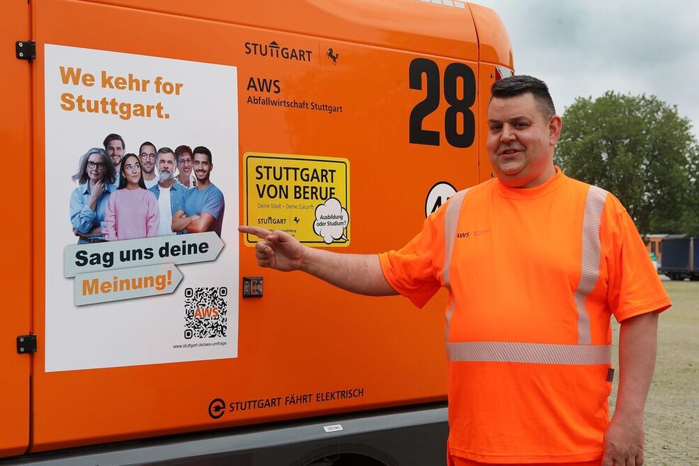 Ein Mitarbeiter der AWS steht in orangefarbener Arbeitskleidung vor einem Fahrzeug der Aws und zeigt auf ein aktuelles Plakat mit dem Hinweis auf die Umfrage.