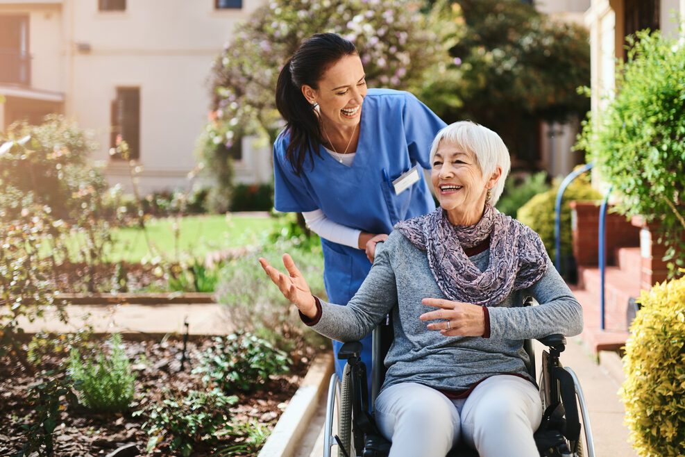 Eine ältere Frau in einem Rollstuhl und eine Pflegerin verbringen in einem Park gemeinsam Zeit miteinander. Im Hintergrund sind eine Wiese und gepflasterte Wege zu erkennen.