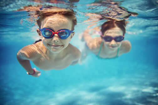 Zwei Kinder mit Taucherbrillen tauchen unter Wasser in einem Schwimmbad.