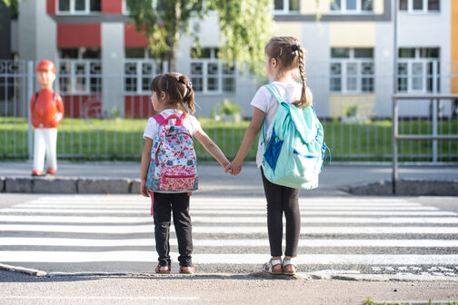Zwei kleine Mädchen stehen an der Straße vor einem Zebrastreifen und halten sich an der Hand.