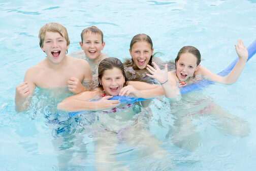 Fünf Kinder schwimmen in einem Schwimmbecken und lachen in die Kamera.
