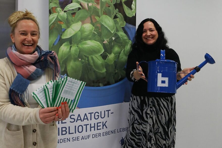 Stefanie Schilling mit Saatgutpäckchen und Alexandra Kirchner mit Bibliotheks-Gießkanne stehen lachend vor einem Plakat zur Saatgutbibliothek