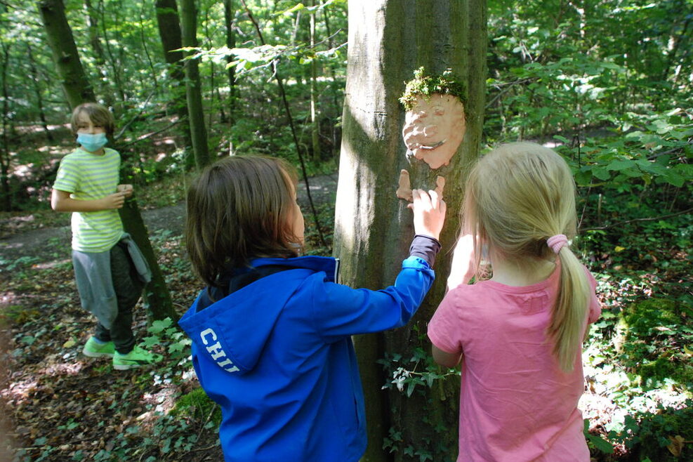 Kinder spielen im Wald. An einem Baumstamm haben sie ein Gesicht aus Knete angebracht.