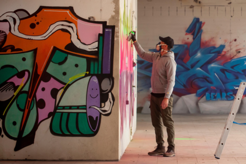 Ein Sprayer srüht ein Graffiti auf eine Wand.