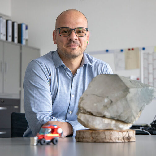Daniel Benneweg sitz an seinem Schreibtisch, im Vordergrund ist ein grauer Stein und ein Spielzeugauto zu sehen.