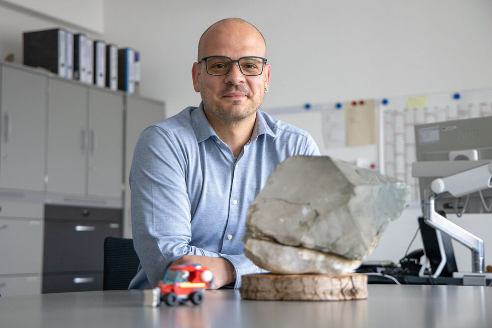 Daniel Benneweg sitz an seinem Schreibtisch, im Vordergrund ist ein grauer Stein und ein Spielzeugauto zu sehen.