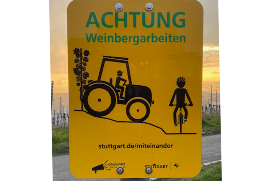 Eines der neuen Hinweisschilder. Auf gelben Hintergrund ist ein gezeichneter Traktor zu sehen der sich einem Fahrradfahrer nähert. Hinter dem Traktor ist eine Weinrebe zu erkennen. Im oberen Bereich des Schildes steht in grüner Schrift: "Achtung Weinbergarbeiten". Im unteren Bereich: "stuttgart.de/miteinander.