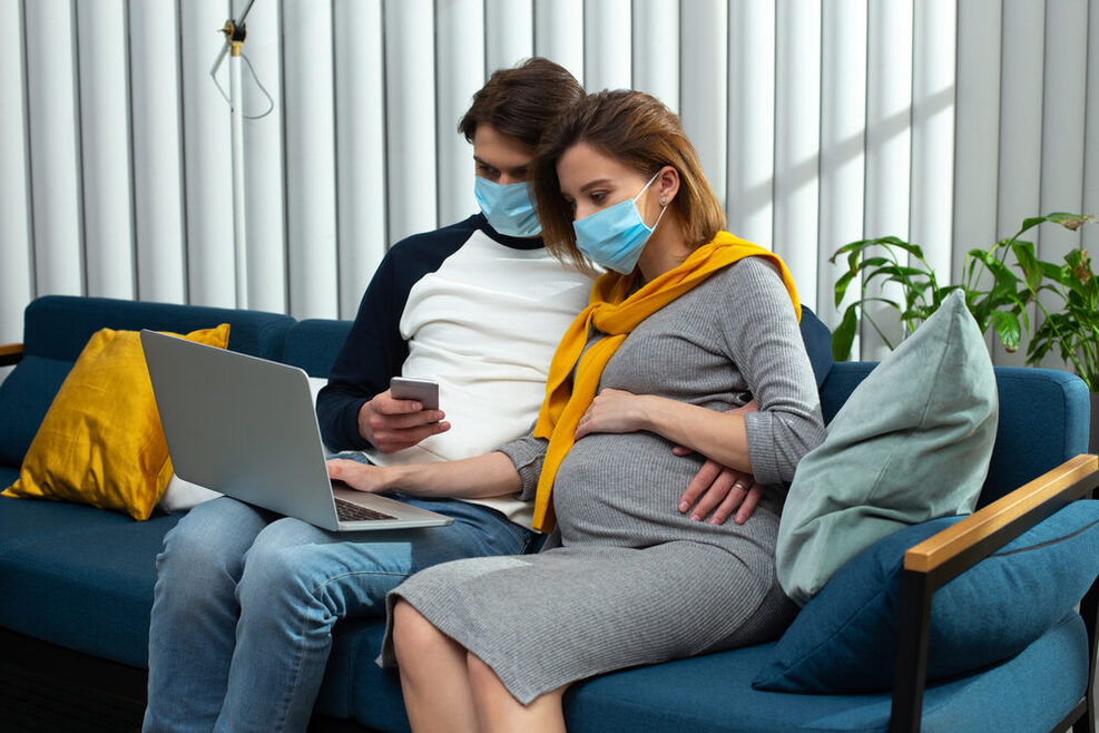 Ein Mann und seine schwangere Frau sitzen auf einem Sofa mit Mundschutz und haben ein Mobiltelefon in der Hand sowie einen aufgeklappten Laptop auf dem Schoß.