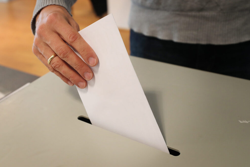Blick auf eine Wahlurne. Eine Person wirft gerade ihren Stimmenzettel in die Urne.