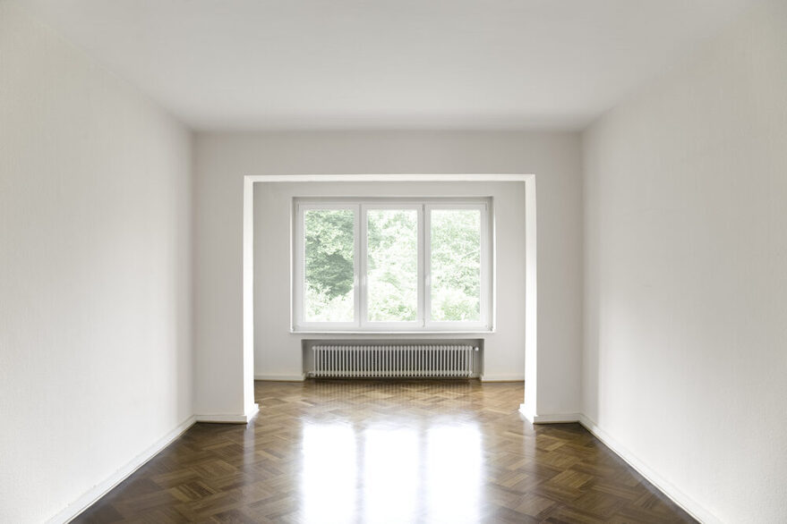 Leerstehendes Zimmer in einer Wohnung mit Parkettboden und Fensterfront am Ende des Raums..