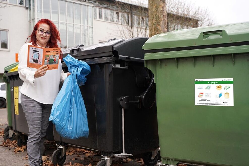 Eine Frau steht vor großen Mülltonnen und hält einen blauen Müllbeutel in der linken Hand. in der rechten hält sie die Abfallbroschüre der AWS.
