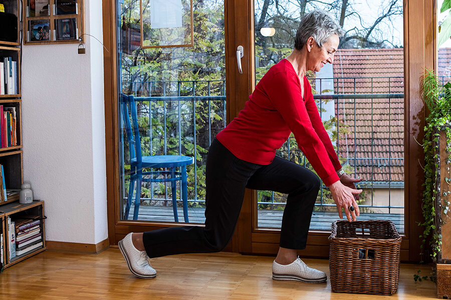 Eine ältere Frau geht bei sich zu Hause im Wohnzimmer in die Hocke, um einen Korb mit geradem Rücken zu heben. Im Hintergrund ist durch das bodentiefe Fenster ein Balkon zu sehen, auf dem ein blauer Stuhl steht.