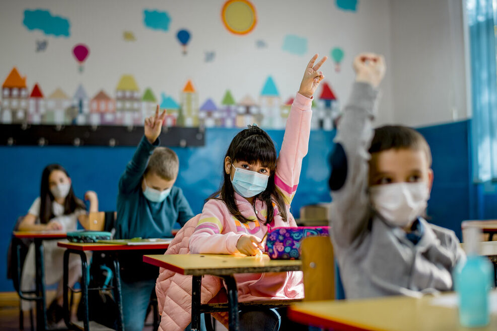 Grundschüler sitzen in einem Klassenraum und tragen eine Maske. Einige melden sich. Im Hintergrund ist eine buntbemalte Wand des Klassenzimmers zu sehen.