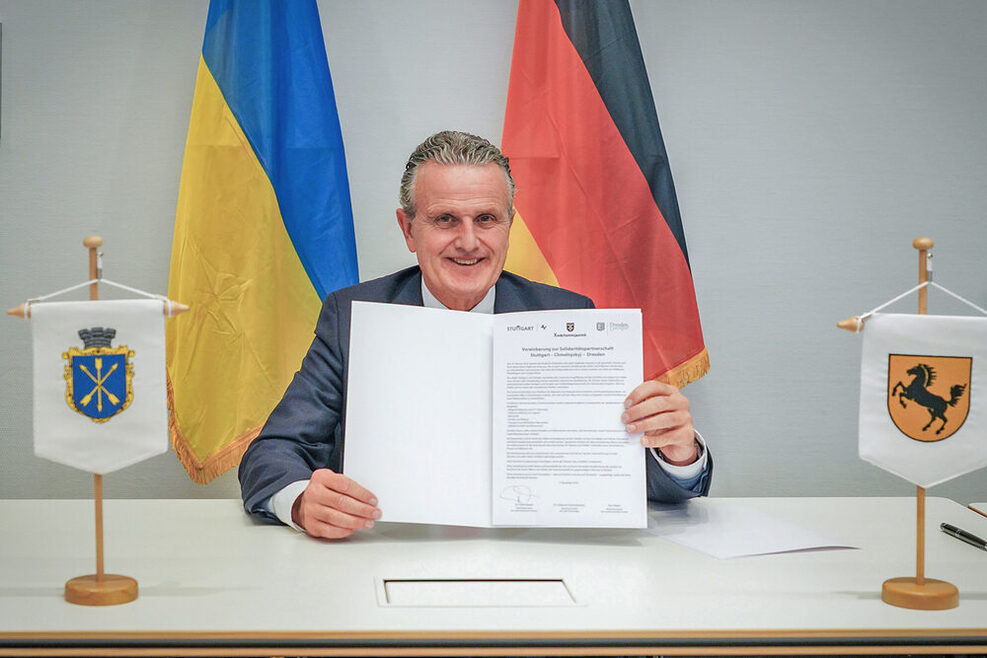 Ein am Schreibtisch sitzender Mann hält ein Schriftstück in die Kamera. Im Hintergrund die ukrainische und die deutsche Flagge, im Vordergrund Wimpel mit den Stadtwappen von Chmelnyzkyj und Stuttgart.