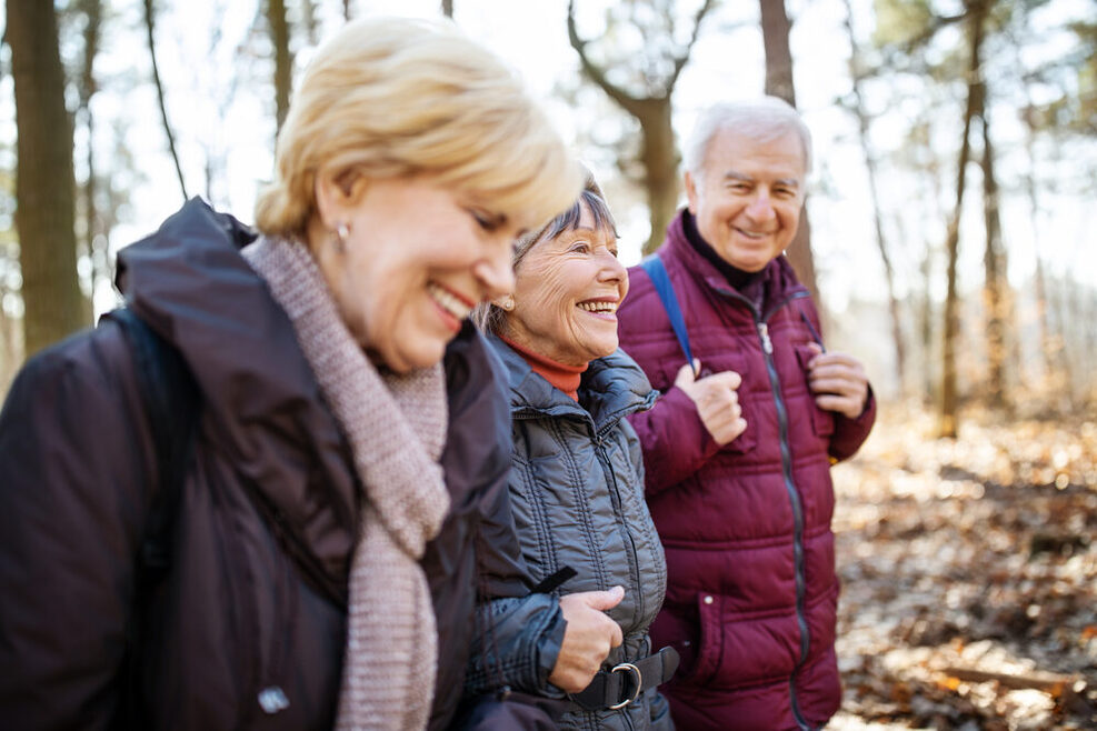 Zwei Seniorinnen und ein Senior gehen in Winterkleidung im Wald spazieren und lachen