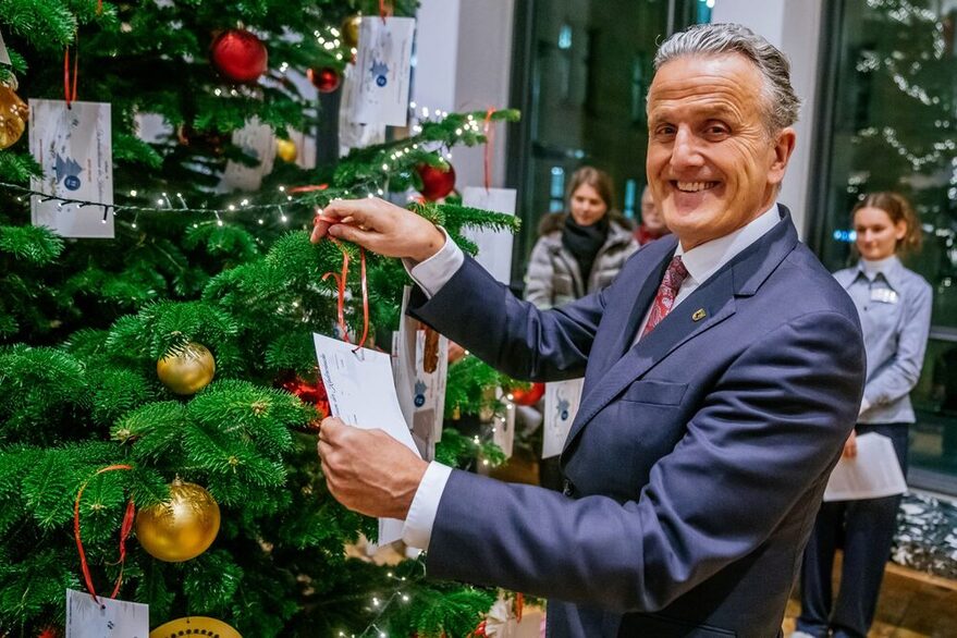 OB Nopper steht vor einem prächtigen Weihnachtsbaum mit grünen Nadeln und golden schimmernden Christbaumkugeln. In seiner Hand hält er eine Karte, die einen Kinderwunsch übermittelt.