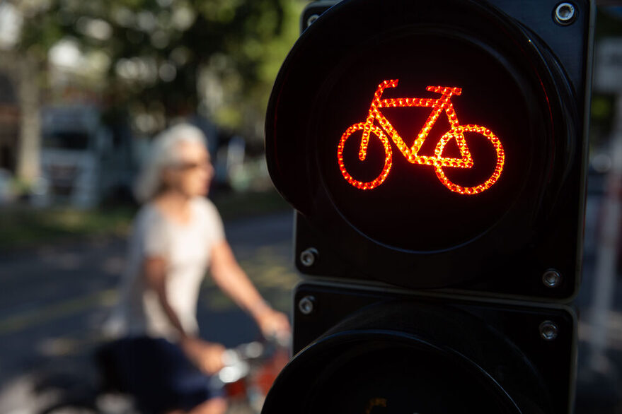 Im Vordergrund ist rechts eine Fahrradampel auf Rot zu sehen. Im Hintergund ist verschwommen eine Radlerin zu erkennen