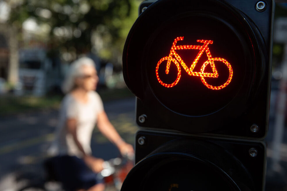 Im Vordergrund ist rechts eine Fahrradampel auf Rot zu sehen. Im Hintergund ist verschwommen eine Radlerin zu erkennen