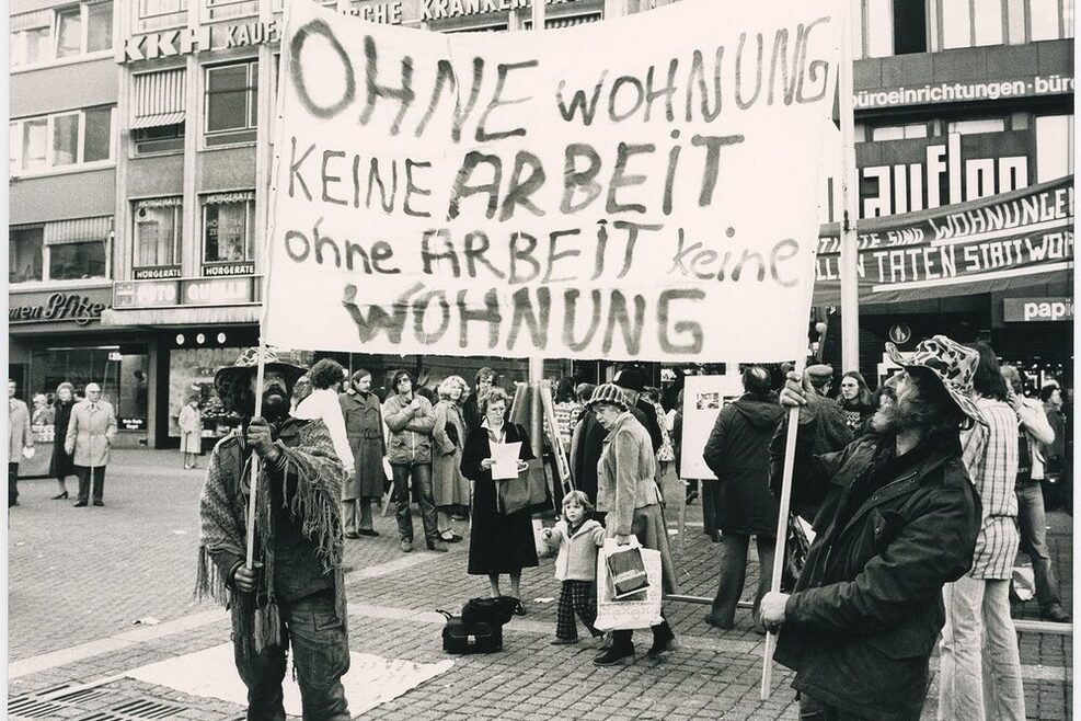 Schwarz-weiß Foto mit zwei Wohnungslosen, die ein Schild hochalten mit der Aufschrift: Ohne WOhnung keine Arbeit, ohne Arbeit keine Wohnung