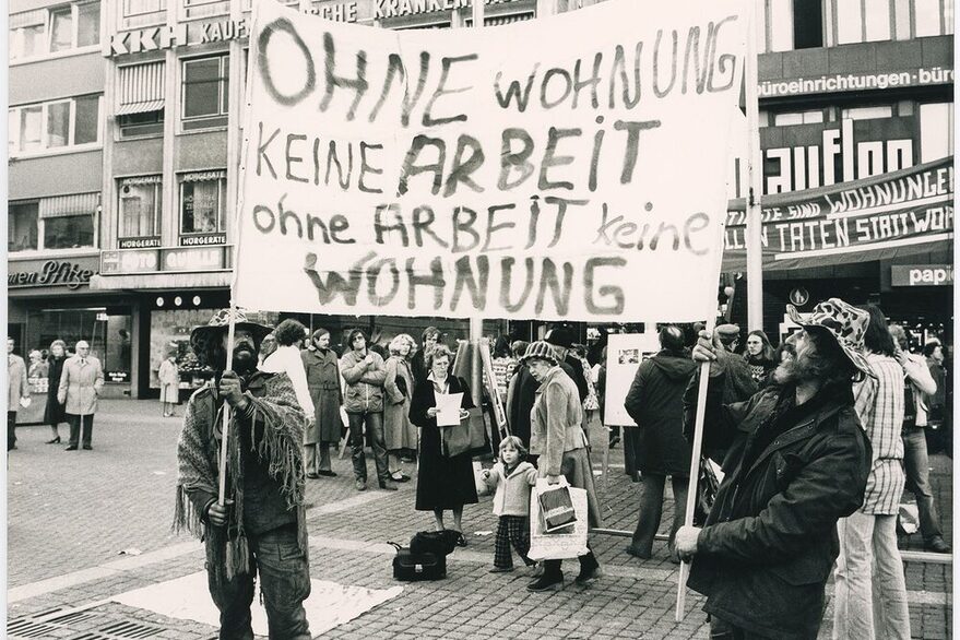 Schwarz-weiß Foto mit zwei Wohnungslosen, die ein Schild hochalten mit der Aufschrift: Ohne WOhnung keine Arbeit, ohne Arbeit keine Wohnung