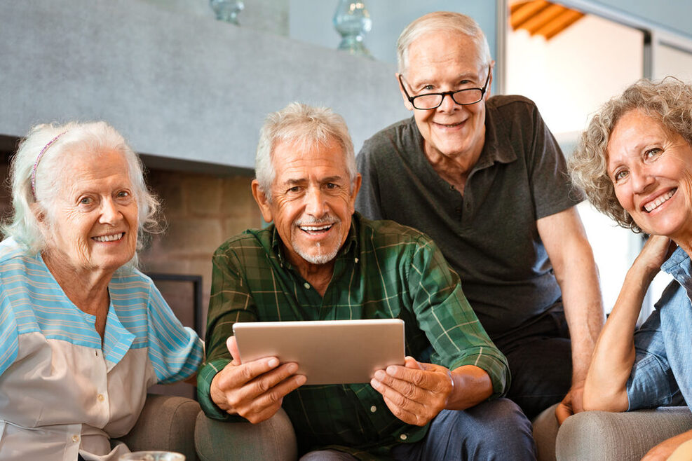 Seniorinnen und Senioren sitzen auf einem Sofa bzw. stehen daneben. Ein Senior hält danei ein Tablet in den Händen.