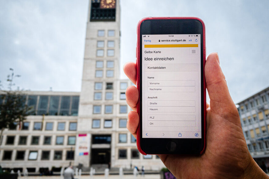 Auf einem Handybildschirm ist die Anwenundg der Gelben Karte zu sehen, im Hintergrund das Stuttgarter Rathaus