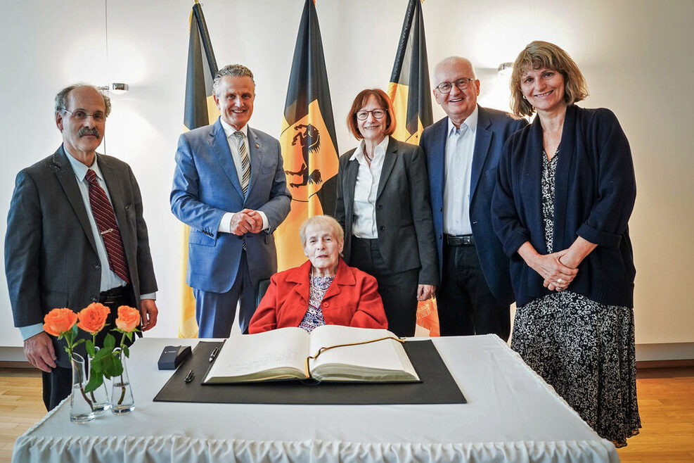 Eine Frau sitzt vor dem Goldenen Buch der Stadt Stuttgart. Im Hintergrund stehen drei Männer und zwei Frauen.