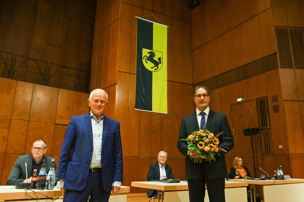 Oberbürgermeister Fritz Kuhn (links) gratuliert dem zukünftigen Bürgermeister für Sicherheit, Ordnung und Sport, Dr. Clemens Maier zur Wahl. Der neue Bürgermeister freut sich, er hält einen Blumenstrauß in der Hand.
