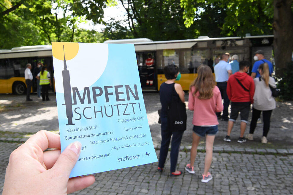 Flyer mit Aufschrift "Impfen schützt" im hintergrund stehen Menschen in einer Schlange vor dem Impfbus.