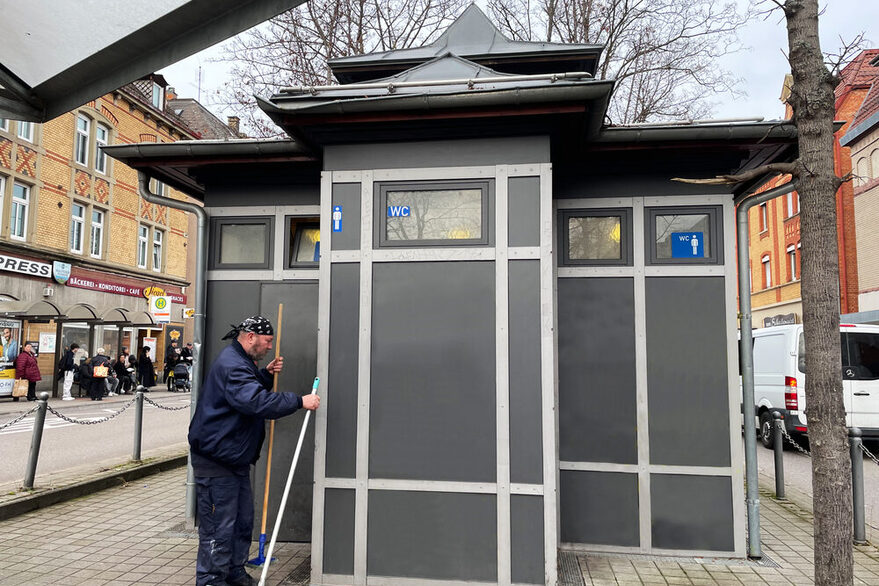 Mann mit zwei Besen in der Hand steht vor einer öffentlichen Toilettenanlage.