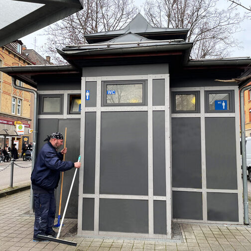Mann mit zwei Besen in der Hand steht vor einer öffentlichen Toilettenanlage.